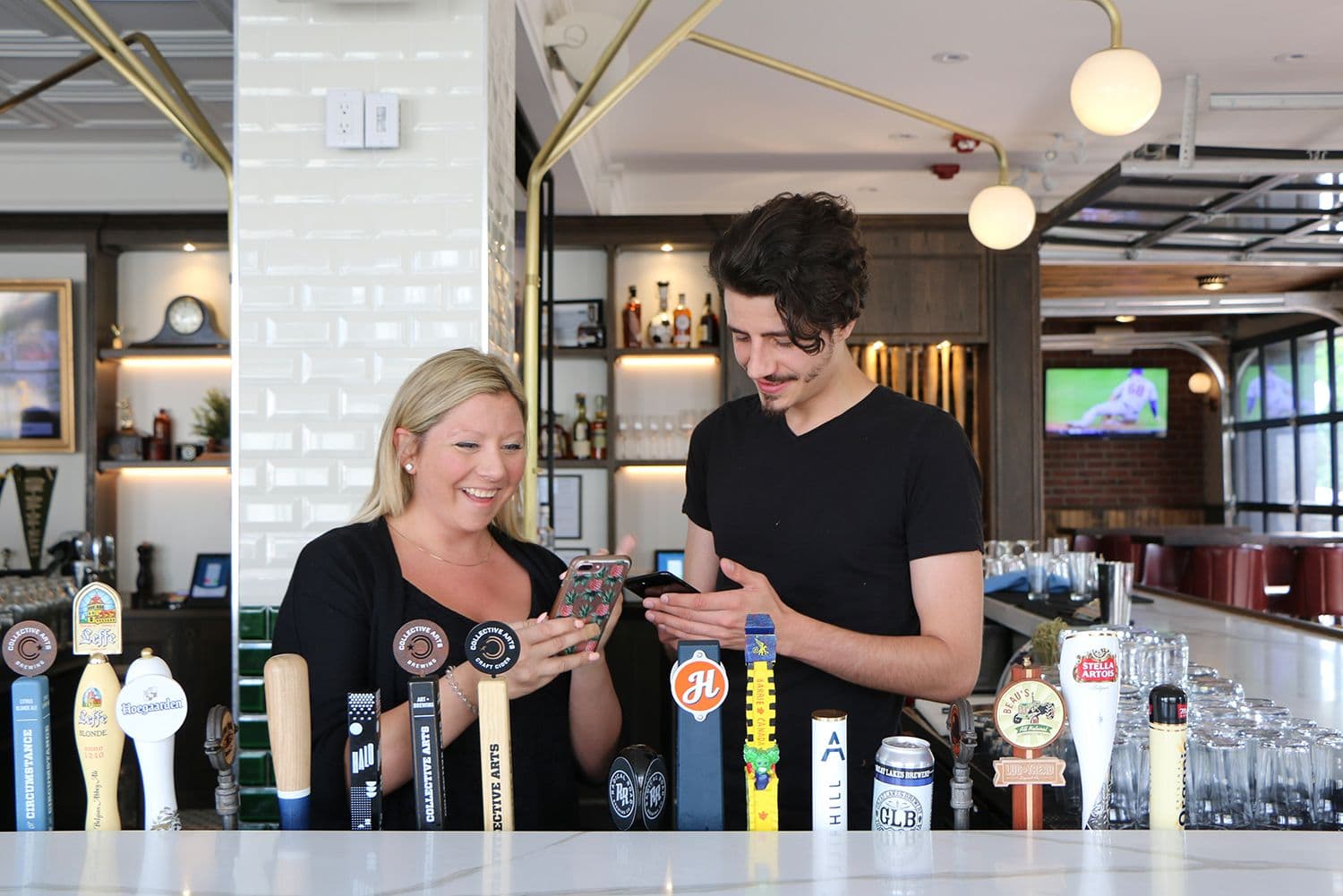 Bartenders using phones behind bar with tap handles.